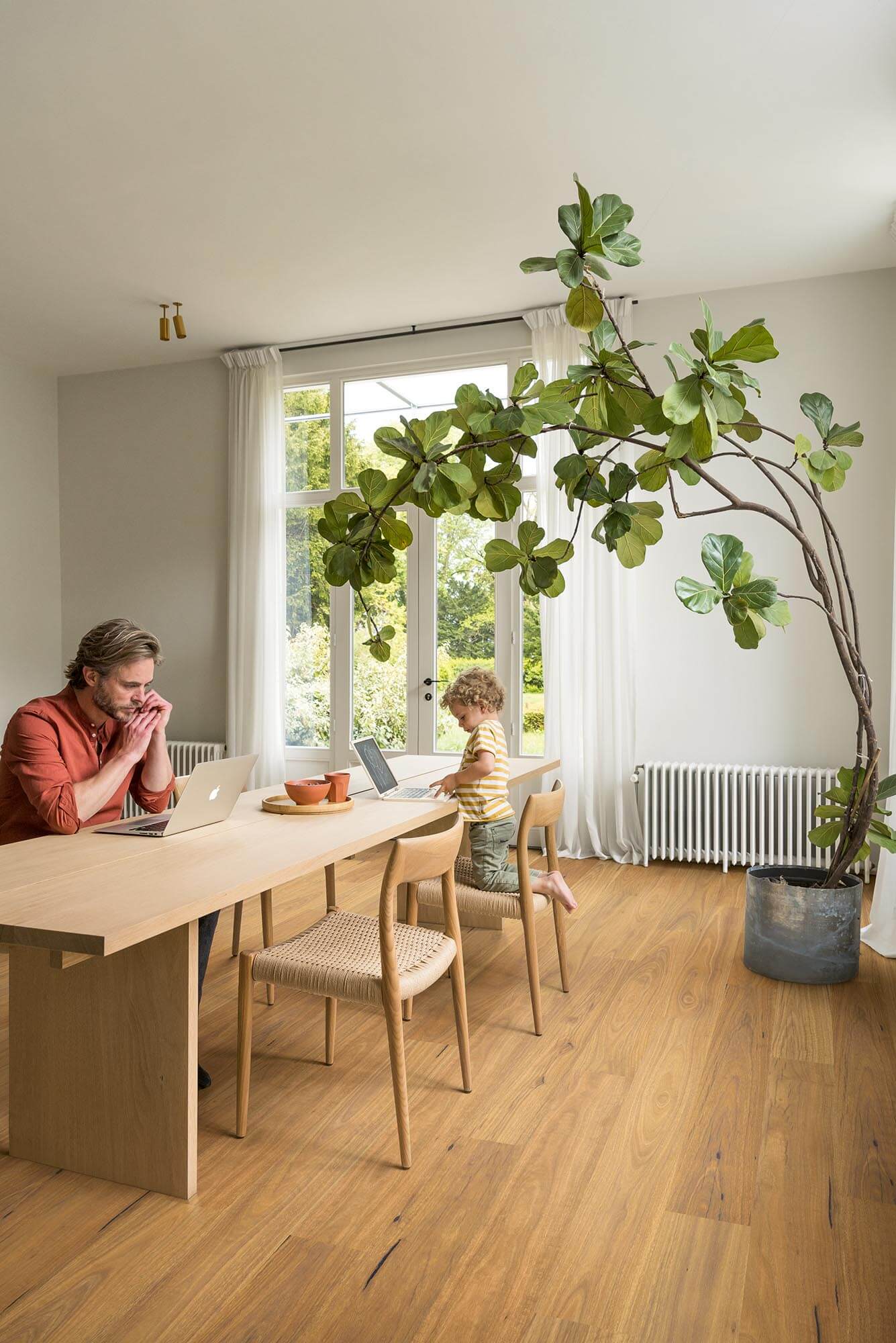 Person working on a laptop at a wooden table with a child, large indoor plant, and bright window in a modern room.
