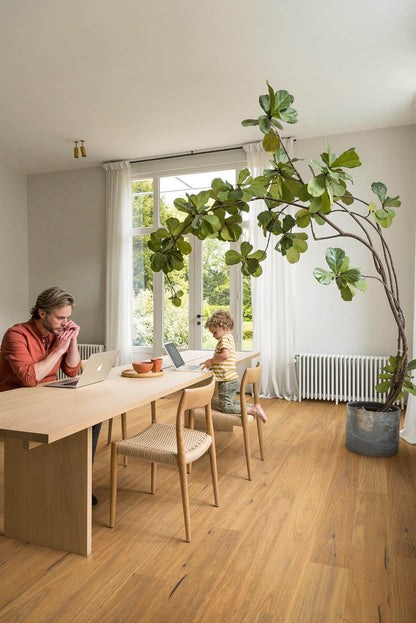 Person working on a laptop at a wooden table with a child, large indoor plant, and bright window in a modern room.